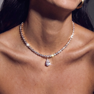 A close-up lifestyle shot of a woman wearing a sparkling diamond necklace with a pear-cut pendant and gold hoop earrings, captured against a dark gray background. | Model Image